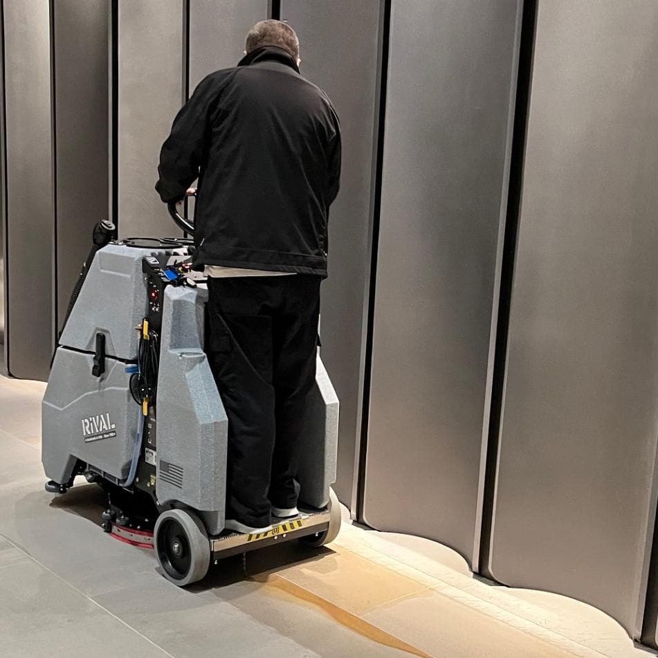 A Tomcat Rival stand on floor scrubber easily cleans the porcelain tile in the hallway of an office building.
